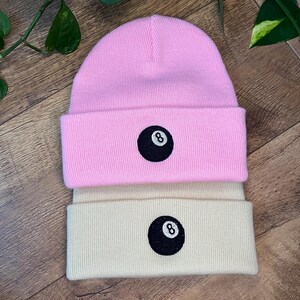 Puede incluir: Dos gorros de punto, uno rosa y otro crema, apilados. Cada gorro presenta un círculo negro con el número 8 bordado en la parte delantera. Los gorros están hechos de un material de punto acanalado suave.