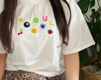 Op de afbeelding: Wit T-shirt met verschillende geborduurde patches, waaronder een ster, een klavertjevier, een hoefijzer, een halve maan, een smiley, een lieveheersbeestje, een blauwe ster en een zwarte biljartbal.