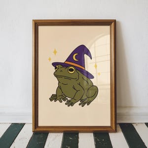 May include: A framed print of a cartoon frog wearing a purple and orange witch's hat with a crescent moon. The frog is green with yellow eyes, and the background is a light beige colour with yellow star accents. The frame is brown.