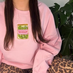 May include: A pink sweatshirt featuring an embroidered design of a jar of pickles. The jar has a pink label with the word "PICKLES" on it.