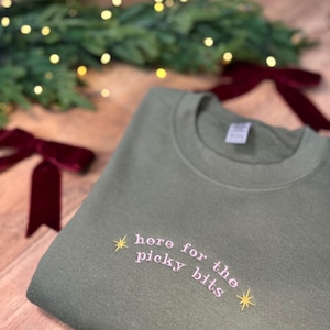 Könnte beinhalten: Ein olivgrünes Sweatshirt mit dem gestickten Text "here for the picky bits" in einer kursiven Schriftart. Der Text ist hellrosa, mit einem Stern auf jeder Seite. Das Sweatshirt ist auf einer Holzoberfläche gefaltet.