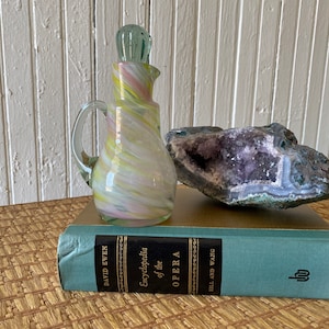 Puede incluir: Una jarra de vidrio blanco, amarillo y rosa con un tapón de vidrio verde se encuentra sobre un libro azul titulado "Enciclopedia de la Ópera" de David Ewen. Un gran geodo de amatista se encuentra detrás de la jarra.
