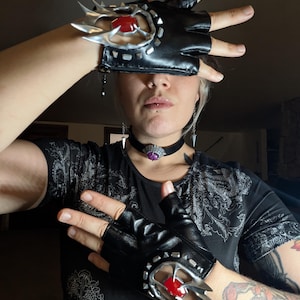 May include: Black fingerless gloves with silver-colored, spiky accents and a red gem. The gloves have a decorative design around the wrist. A matching choker with a purple gem is also visible.