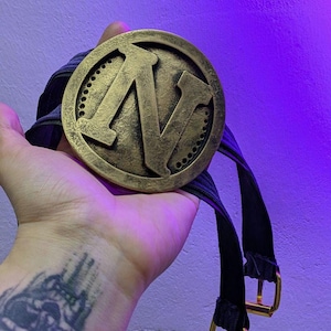 May include: A circular gold-coloured medal featuring the letter 'N' in the centre. It is attached to a black ribbon with gold-coloured buckles. The medal has a worn, vintage appearance, suggesting it is an award or commemorative item.