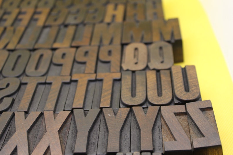 May include: A close-up of a set of wooden letterpress printing blocks, arranged in alphabetical order from A to Z. The blocks are made of dark brown wood and have a worn, vintage look.