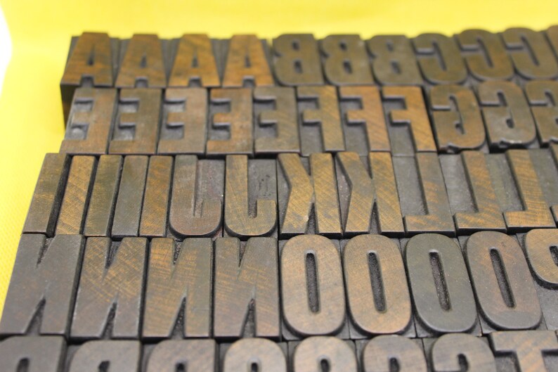 May include: A collection of vintage wooden letterpress printing blocks. The blocks are made of dark wood and have a worn, antique look. The letters include A, B, C, D, E, F, G, H, I, J, K, L, M, N, O, P, Q, R, S, T, U, V, W, X, Y, Z, 0, 1, 2, 3, 4, 5, 6, 7, 8, 9.