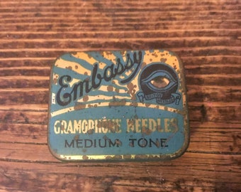 May include: A vintage tin box for gramophone needles. The box is blue and yellow with the words "Embassy Gramophone Needles Medium Tone" printed on it. The box has a logo of an eye with the words "British Needles" printed on a banner.