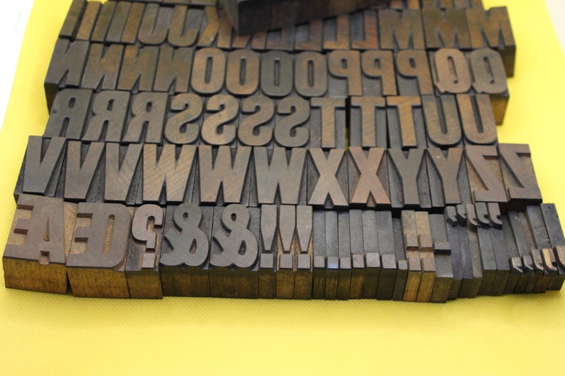 May include: A collection of wooden letterpress printing blocks, including uppercase letters, numbers, punctuation marks, and symbols. The blocks are arranged in rows and are made of dark brown wood.