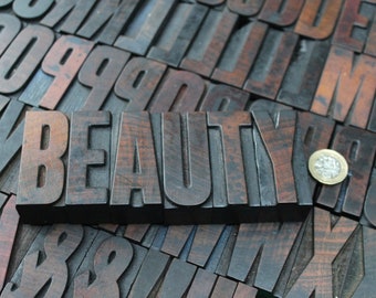 May include: Wooden letterpress blocks spelling the word "BEAUTY" in a bold font. The letters are dark brown and sit atop a jumble of other letterpress blocks. A coin rests near the word.