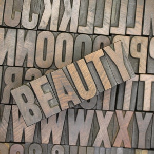 May include: The word "BEAUTY" spelled out in wooden letterpress blocks. The blocks are arranged in a row and are made of dark brown wood with a grain pattern. The letters are raised and have a slightly worn appearance.