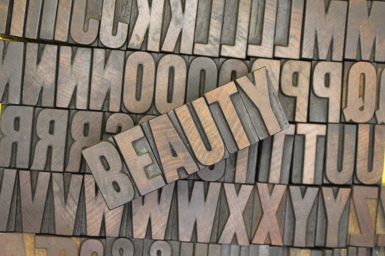 May include: The word "BEAUTY" spelled out in wooden letterpress blocks. The blocks are arranged in a row and are made of dark brown wood with a grain pattern. The letters are raised and have a slightly worn appearance.