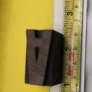 May include: A dark brown wooden letter V, measuring approximately 2 inches in height, is placed on a yellow surface next to a yellow tape measure. The tape measure reads "Stanley" and "Made in England".