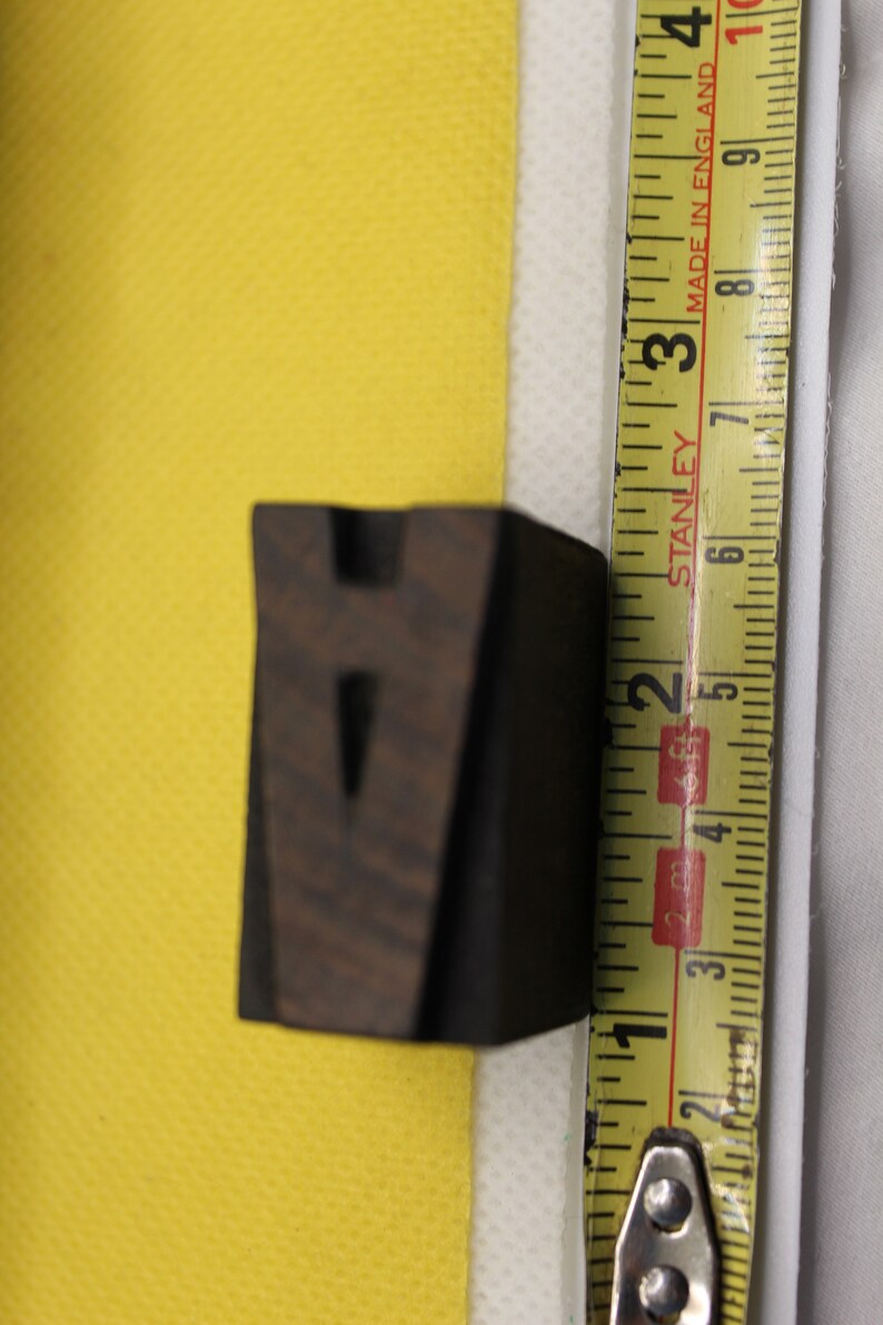 May include: A dark brown wooden letter V, measuring approximately 2 inches in height, is placed on a yellow surface next to a yellow tape measure. The tape measure reads "Stanley" and "Made in England".