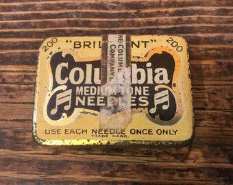 May include: A yellow metal tin with black lettering that reads "Columbia Medium Tone Needles" and "Use Each Needle Once Only." The tin has a musical note on each side and the words "200 "Brilliant" 200" on the top.