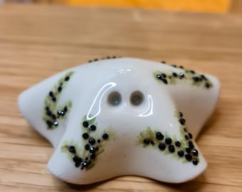 May include: A white ceramic starfish-shaped salt and pepper shaker featuring two small holes on the top centre and a hand-painted green leafy vine pattern with small black dots accenting the five points of the star-shaped object.