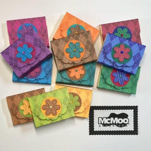 May include: A collection of colorful, handmade paper gift boxes. Each box features a diamond pattern and a decorative flower with a gem center. The boxes are in various colors, including purple, green, and brown. The McMoo Creations logo is also visible.