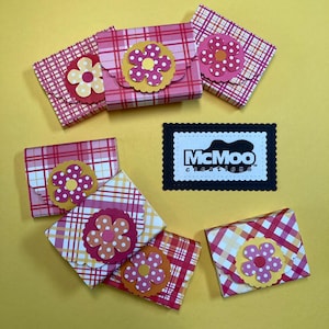 May include: A collection of small, plaid-patterned gift boxes in shades of pink and red, each adorned with a layered flower embellishment. The boxes are arranged on a yellow surface, with a black and white logo that reads "McMoo creations".