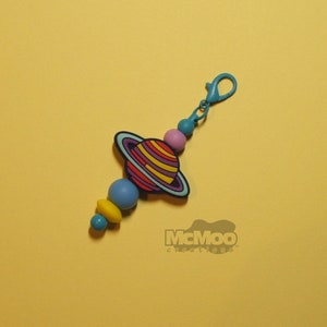 May include: A colorful beaded keychain with a blue, yellow, and pink bead design. The keychain features a charm of a cartoon planet with a ring around it. The keychain has a blue metal clasp.