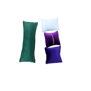 Peut inclure: Un assortiment de petits coussins décoratifs de différentes couleurs. Les coussins comprennent un long traversin vert foncé, un coussin rectangulaire blanc, un coussin carré violet avec une ligne dorée et un coussin carré violet foncé.