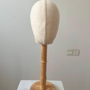 May include: A beige fabric head mannequin on a wooden stand. The head form is oval-shaped and made of a light-colored fabric. The wooden stand has a round base and a vertical pole, all in a natural wood finish. The mannequin is designed for displaying wigs or hats.