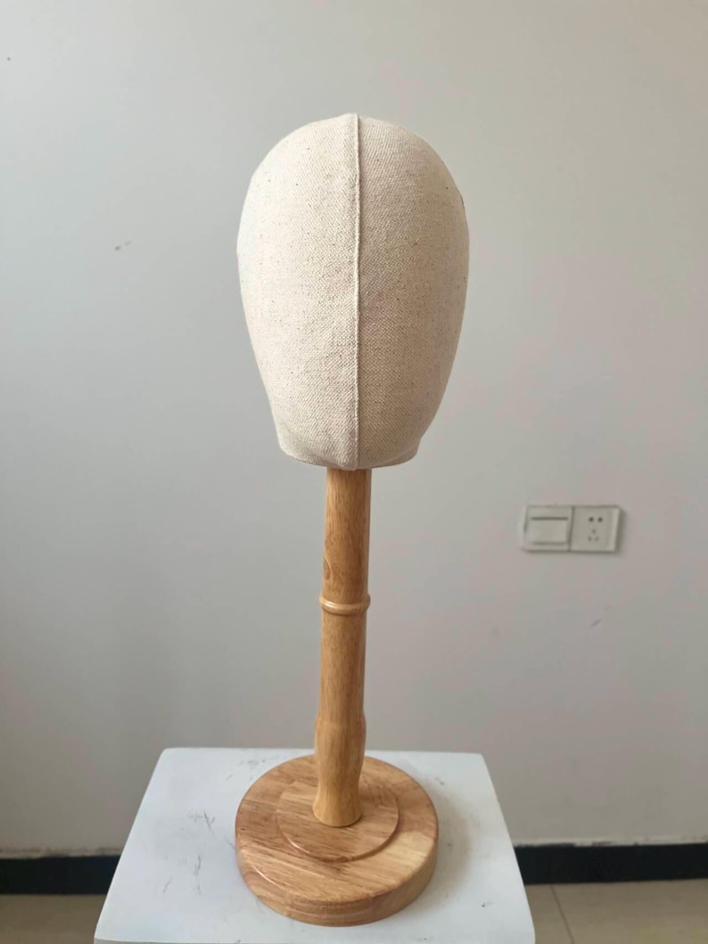 May include: A beige fabric head mannequin on a wooden stand. The head form is oval-shaped and made of a light-colored fabric. The wooden stand has a round base and a vertical pole, all in a natural wood finish. The mannequin is designed for displaying wigs or hats.