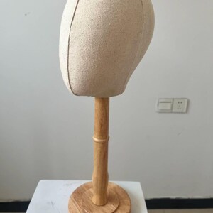May include: A beige fabric mannequin head on a wooden stand. The head is oval-shaped with a seam down the middle. The stand has a round base and a cylindrical pole, all made of light-colored wood.