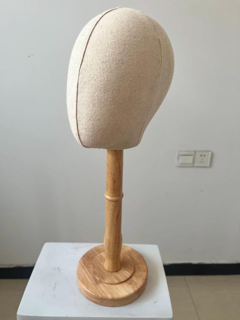 May include: A beige fabric mannequin head on a wooden stand. The head is oval-shaped with a seam down the middle. The stand has a round base and a cylindrical pole, all made of light-colored wood.