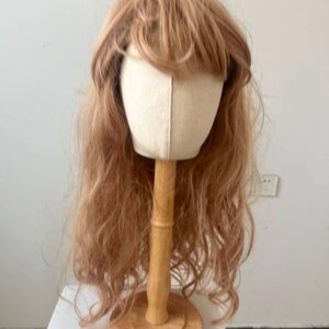 May include: A light brown wig with long, wavy hair and bangs is displayed on a canvas head form. The head form is mounted on a wooden stand with a round base. The wig is styled to appear natural.