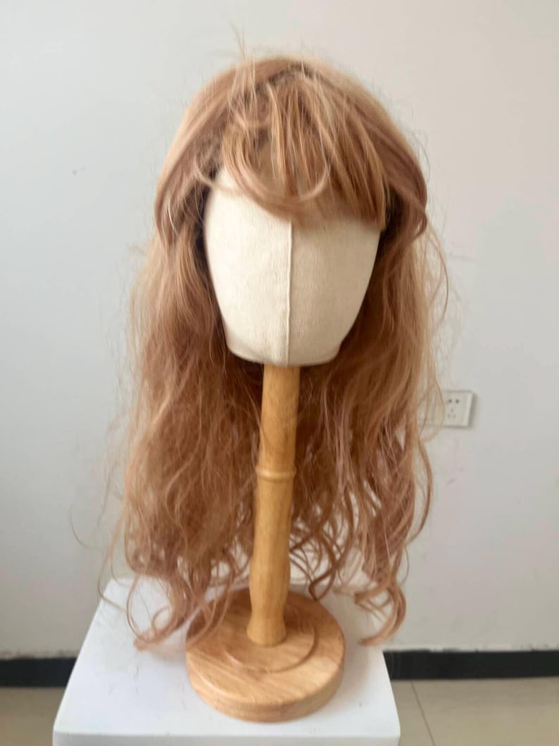 May include: A light brown wig with long, wavy hair and bangs is displayed on a canvas head form. The head form is mounted on a wooden stand with a round base. The wig is styled to appear natural.