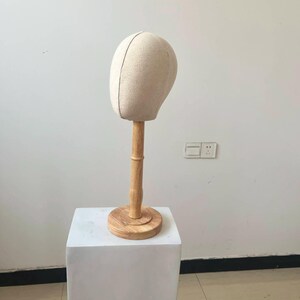 May include: A beige fabric wig head on a wooden stand. The head is oval-shaped with a vertical seam down the center. The stand has a round base and a cylindrical neck, all made of light-colored wood. The head is placed on a white square pedestal.