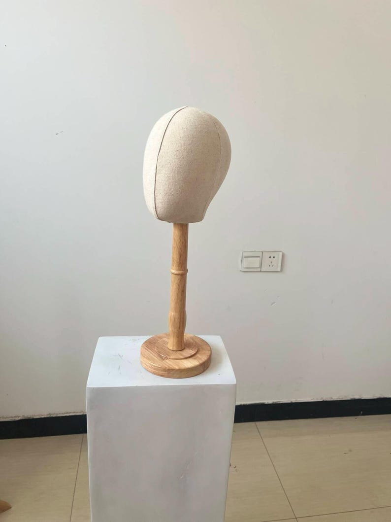 May include: A beige fabric wig head on a wooden stand. The head is oval-shaped with a vertical seam down the center. The stand has a round base and a cylindrical neck, all made of light-colored wood. The head is placed on a white square pedestal.
