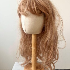 May include: A light brown wig with bangs is displayed on a canvas head form attached to a wooden stand. The wig has long, wavy hair and is styled to appear natural. The wooden stand has a round base.