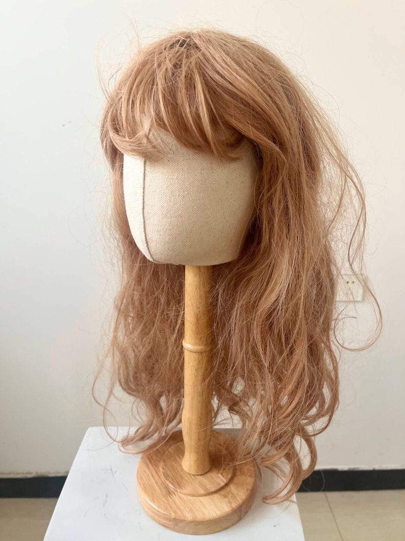 May include: A light brown wig with bangs is displayed on a canvas head form attached to a wooden stand. The wig has long, wavy hair and is styled to appear natural. The wooden stand has a round base.
