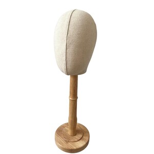 May include: A beige fabric head form on a wooden stand. The head form is oval-shaped and has a seam down the middle. The wooden stand has a round base and a vertical support with a bamboo-like design. The head form is used for displaying hats and wigs.