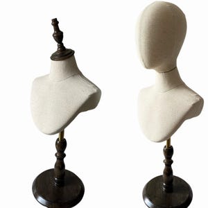 May include: Two dressmaker's bust forms covered in fabric. They are mounted on dark wooden stands with a round base and a brass-coloured support rod. One form has a head and the other has a neck and shoulder form.