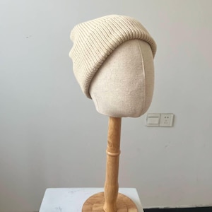 May include: A cream-colored knit beanie hat displayed on a mannequin head. The hat has a ribbed texture and a folded brim. The mannequin head is a light beige color and is mounted on a wooden stand with a round base. The background is a plain white wall.