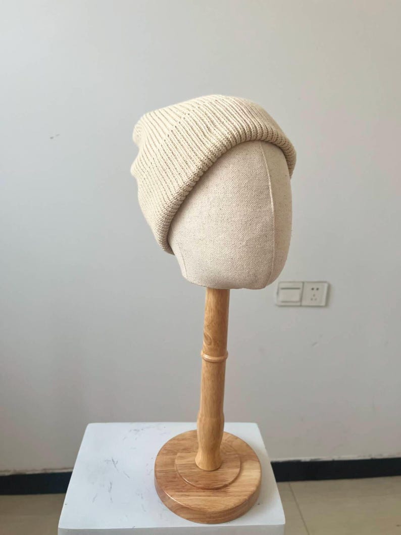May include: A cream-colored knit beanie hat displayed on a mannequin head. The hat has a ribbed texture and a folded brim. The mannequin head is a light beige color and is mounted on a wooden stand with a round base. The background is a plain white wall.