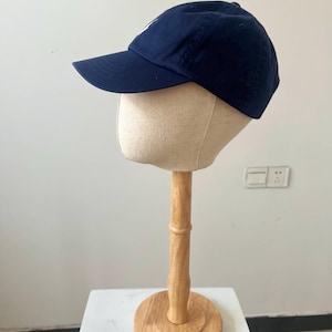 May include: A navy blue baseball cap with a curved brim and a white logo on the front. The cap is displayed on a mannequin head atop a wooden stand with a round base. The stand is on a white surface.