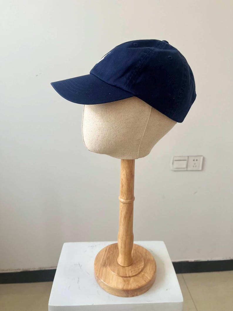 May include: A navy blue baseball cap with a curved brim and a white logo on the front. The cap is displayed on a mannequin head atop a wooden stand with a round base. The stand is on a white surface.