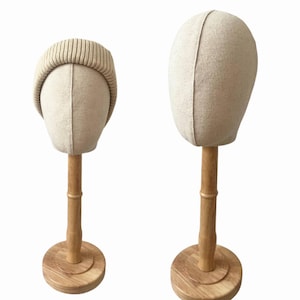 May include: Two beige fabric mannequin heads on wooden stands. One head wears a ribbed knit beanie. The stands have a round wooden base and a bamboo-style pole. The heads are for displaying hats and other headwear.