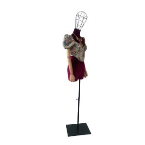 May include: A vintage dress form with a wire head and wooden arms. It displays a floral-print top, a burgundy skirt, and a gold necklace. The form is mounted on a black metal stand, suitable for showcasing clothing.