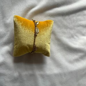 K&ouml;nnte beinhalten: Ein goldenes Kettenarmband mit kleinen Blatt-Anh&auml;ngern auf einem gelben Samtkissen.