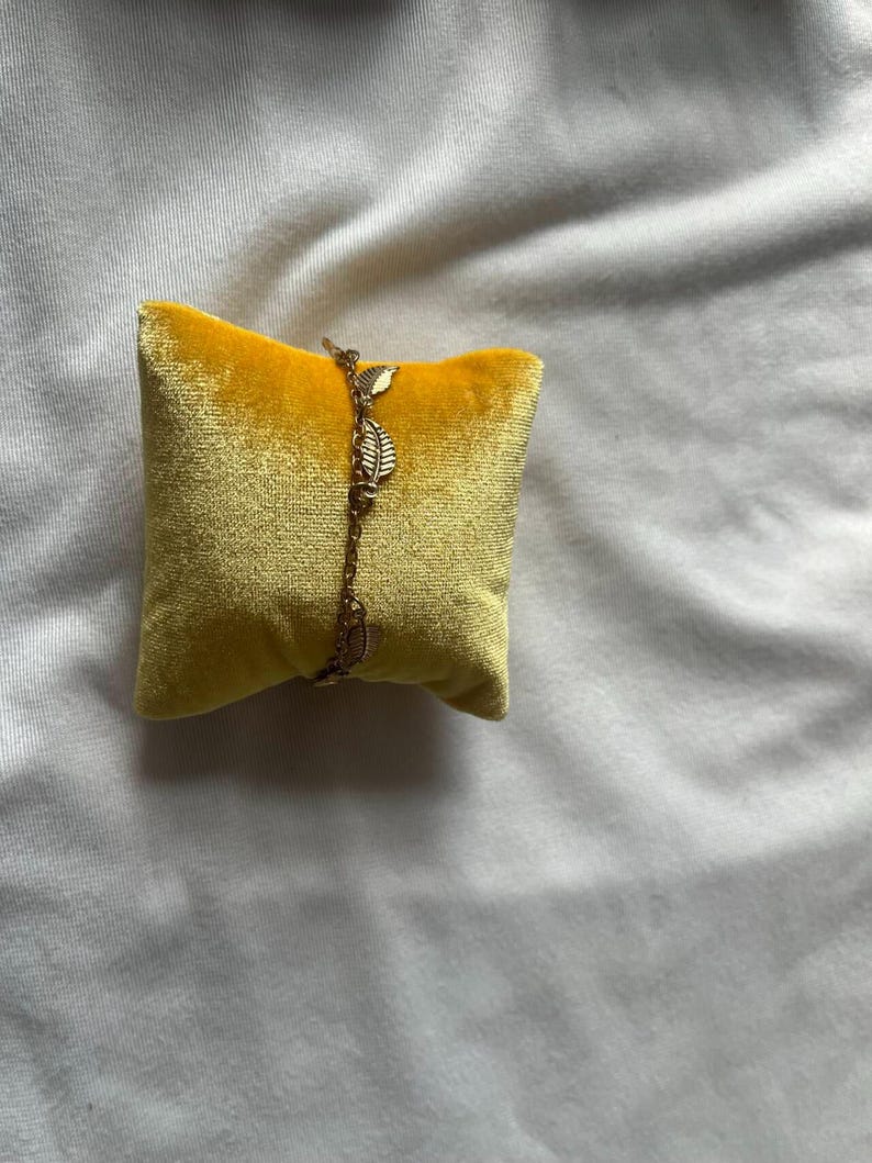 K&ouml;nnte beinhalten: Ein goldenes Kettenarmband mit kleinen Blatt-Anh&auml;ngern auf einem gelben Samtkissen.