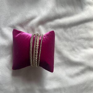 K&ouml;nnte beinhalten: Ein Set aus vier silbernen Armb&auml;ndern mit klaren Strasssteinen, auf einem rosa Samtkissen pr&auml;sentiert.