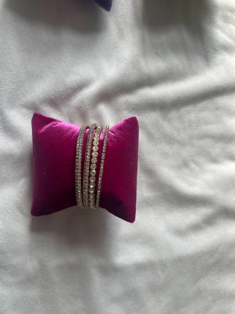 K&ouml;nnte beinhalten: Ein Set aus vier silbernen Armb&auml;ndern mit klaren Strasssteinen, auf einem rosa Samtkissen pr&auml;sentiert.