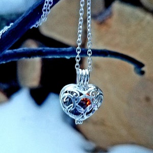 May include: A silver heart-shaped pendant necklace with an intricate filigree design. The pendant holds a small, orange-red gemstone. The necklace hangs from a delicate chain, displayed against a snowy background and a dark blue branch.