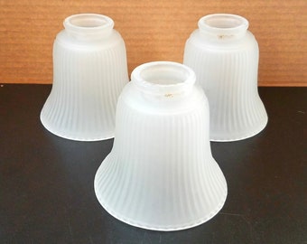 May include: Three frosted glass lamp shades with a bell shape and vertical ribbed design. The shades are white and have a slightly flared bottom. The shades are for a lamp or light fixture.