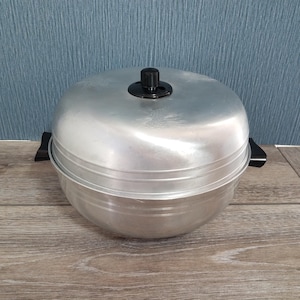 May include: A round, aluminum steamer pot with a black handle and lid knob. The pot has a two-part design with a base and a lid, and is sitting on a wooden surface.
