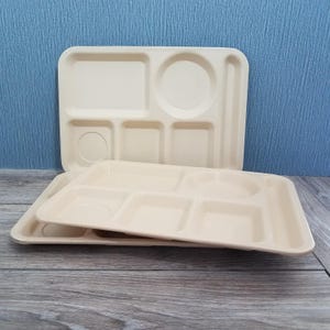 May include: Two beige cafeteria trays with multiple compartments for food. The trays have rectangular, square, and circular sections. One tray is stacked on top of the other, with a blue background.