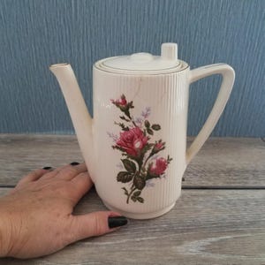 May include: Cream-colored ceramic coffee pot with a lid and a handle. The pot features a vertical ribbed texture and a floral design of pink roses and green leaves. The spout is long and slender.
